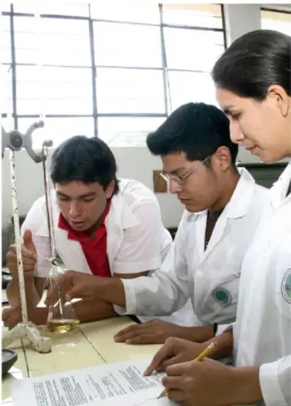 Fotografía de tres estudiantes de secundaria con bata blanca en un laboratorio escolar; uno realiza una titulación con bureta y matraz mientras los otros toman notas en una hoja de trabajo.
