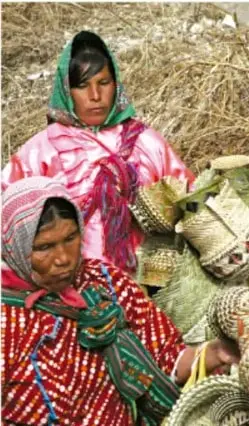 Fotografía de dos mujeres indígenas con ropa tradicional tejida de colores (rosa y rojo) y turbantes, vendiendo cestas y artesanías de palma sobre tierra seca.