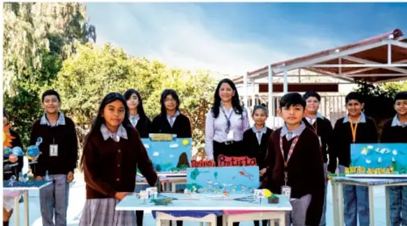 Fotografía de un grupo de estudiantes de telesecundaria con sus maestros presentando maquetas y proyectos escolares en mesas, en un patio o espacio escolar al aire libre con árboles y estructuras de sombra al fondo.