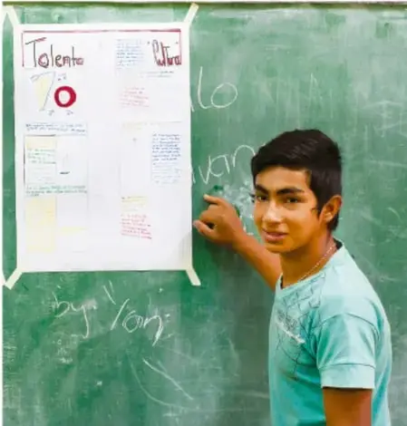 Fotografía de un estudiante de secundaria junto a un cartel de trabajo colocado en el pizarrón de la clase