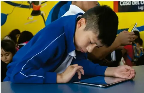 Estudiante con uniforme azul apoyado sobre un pupitre, utilizando una tableta o escritura, en un salón de clases con otros compañeros alrededor; fotografía que ilustra la participación escolar inclusiva.