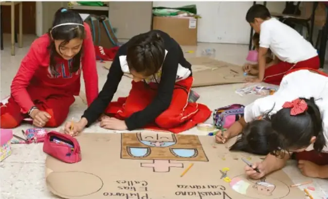 Fotografía de varias estudiantes de primaria trabajando en el piso sobre un mural de cartón. Una niña con suéter rojo escribe, otra con suéter negro dibuja, y dos más observan o colaboran. Hay materiales de arte y un dibujo con figuras y palabras como "Pintores" y "Cuidando".