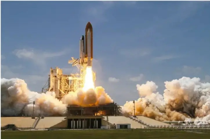 Fotografía de un transbordador espacial despegando desde una plataforma, con una gran nube de humo y fuego en la base, bajo un cielo azul con nubes.