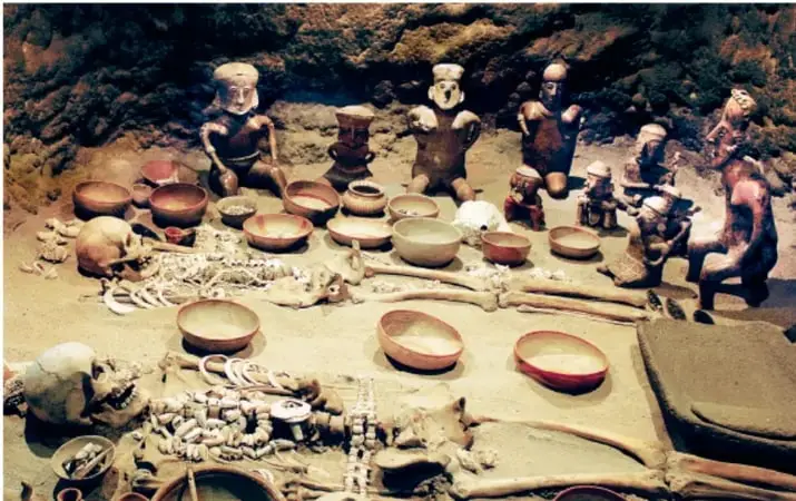 Fotografía de una excavación arqueológica: figuras humanas de barro, vasijas, cuencos, huesos y ajuares dispuestos en el suelo de una tumba o sitio de entierro.