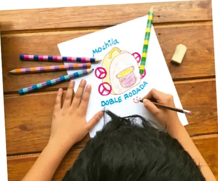 Fotografía de manos dibujando una mochila con ruedas sobre una hoja, con lápices de colores y borrador sobre una mesa de madera.