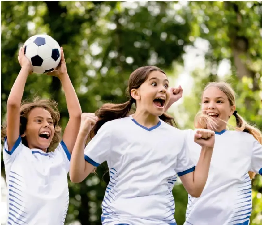 Tres niñas con uniforme deportivo blanco y azul celebran alegremente; una sostiene un balón de fútbol. Están al aire libre con árboles de fondo.