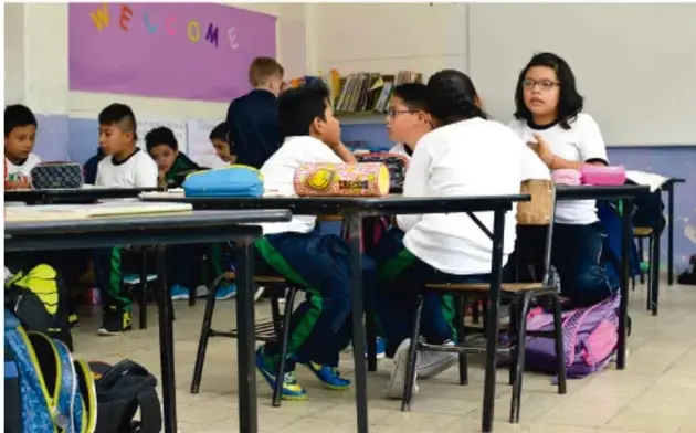Fotografía de estudiantes de primaria sentados en equipos dentro del aula, con mochilas y cuadernos, participando en una actividad colaborativa.