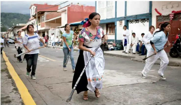 Fotografía de un grupo de personas, principalmente mujeres con vestimenta tradicional, corriendo por una calle con palos largos, en lo que parece ser un juego de pelota tradicional.