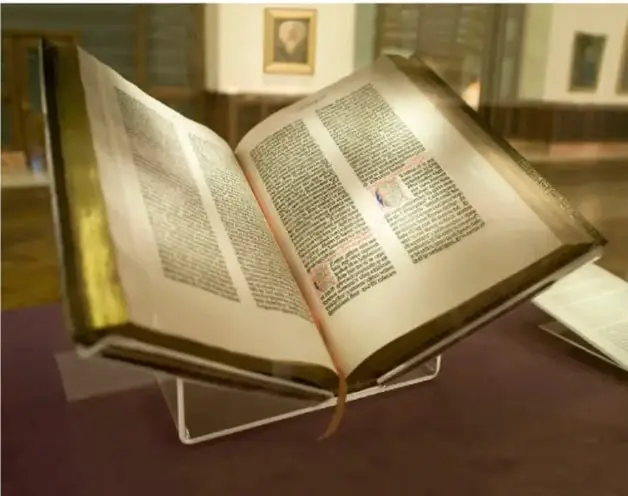 Fotografía de un libro antiguo abierto sobre un soporte de cristal, con páginas de texto impreso en dos columnas y capitulares iluminadas, en lo que parece ser una biblioteca o sala de exposición.