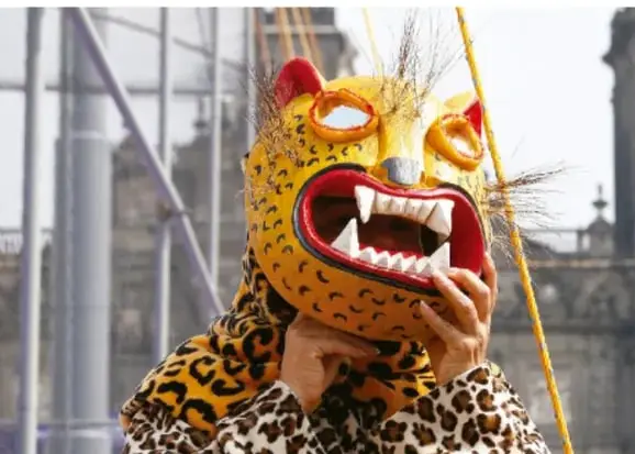 Fotografía de una máscara ritual de felino (jaguar) con colmillos y ojos destacados, sostenida por una persona con traje de piel moteada, probablemente en una ceremonia o manifestación cultural indígena.
