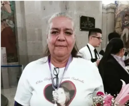 Fotografía de Norma Andrade, mujer de edad avanzada con cabello canoso, usando una camiseta blanca con un corazón y la imagen de una persona. Al fondo, otras personas en un espacio público.