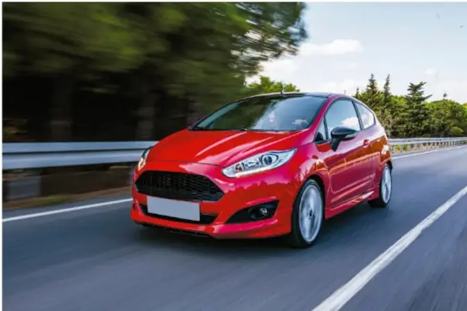 Automóvil rojo en movimiento sobre una carretera, con fondo de árboles desenfocados por la velocidad.