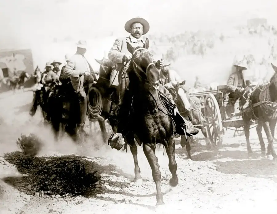 Fotografía histórica en sepia: el general Francisco Villa a caballo, armado, encabezando a sus tropas con carretas y soldados, durante la Toma de Ojinaga, enero de 1914.