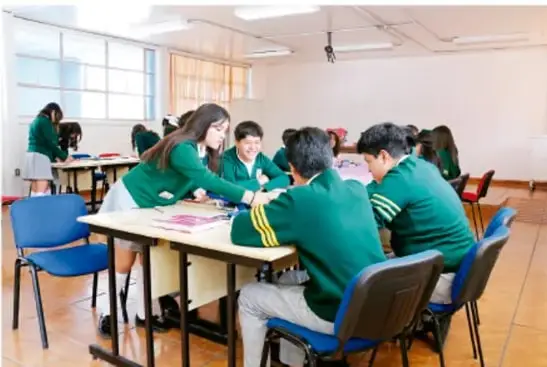 Fotografía de estudiantes de secundaria con uniforme verde, sentados alrededor de una mesa en el aula, trabajando en equipo con papeles de colores para preparar el material de exposición del proyecto.