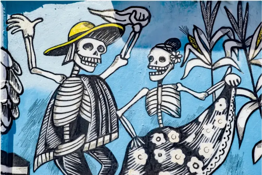 Grafiti de estilo mexicano con calaveras o catrines bailando, con sombreros y vestidos floreados, rodeados de matas de maíz, en azul, blanco y negro.