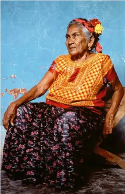 Fotografía de una mujer anciana indígena sentada, con rebozo y falda tradicionales y flores en el cabello, frente a una pared azul; ilustra la participación de las personas mayores en la comunidad.