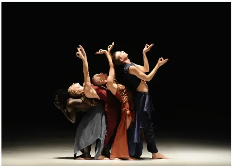 Fotografía escénica de tres bailarines sobre fondo negro: dos mujeres y un hombre con vestuario en tonos gris, rojo y azul oscuro, con los brazos elevados en postura expresiva. Corresponde a la obra Aajej de Virpi Pahkinen, fotografiada por Christa Cowrie.