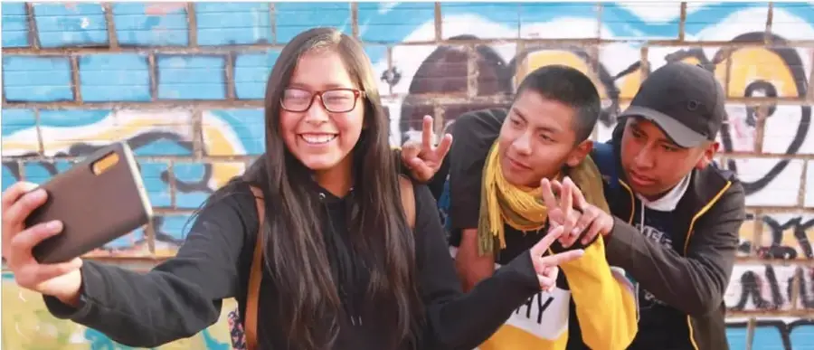 Fotografía de tres jóvenes sonrientes frente a un muro pintado con grafitis de colores; una chica con lentes y mochila toma una selfie con un teléfono celular y dos chicos posan detrás haciendo gestos con las manos.