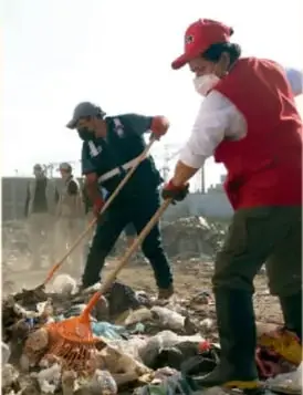 Fotografía de dos personas con chalecos reflectantes y gorras recogiendo basura en un terreno contaminado, como parte de una jornada de limpieza.