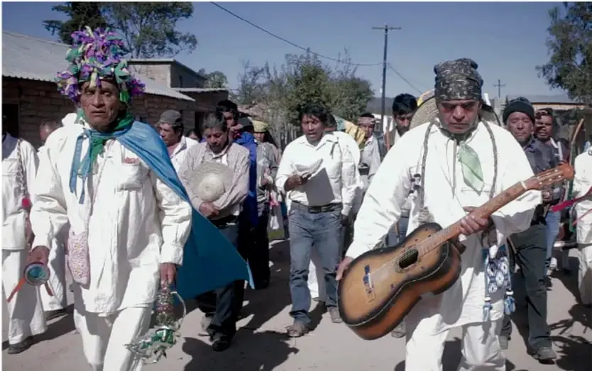 Fotografía de una comunidad indígena mexicana en celebración: hombres con trajes blancos, sombreros adornados con flores y cintas, uno de ellos tocando una guitarra, otros con instrumentos y cohetes, en una calle de una localidad rural.