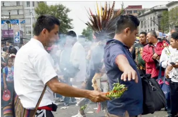 Fotografía de una ceremonia o ritual en la calle: una persona con penacho ofrece frutos o flores a otra entre humo, con público observando, ilustrando los rituales de sanación colectivos.
