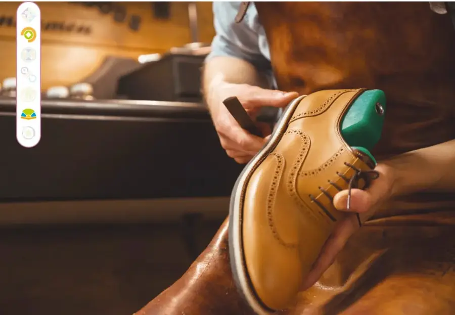 Fotografía de un zapatero con mandil de piel trabajando en su taller: sostiene un zapato de piel color ocre con un formador verde, mientras talla o pule la suela con una herramienta; al fondo se ve una máquina de coser antigua.