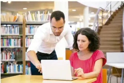 Dos personas (un hombre y una mujer) trabajando con una computadora portátil en una biblioteca con estantes de libros y una escalera al fondo.