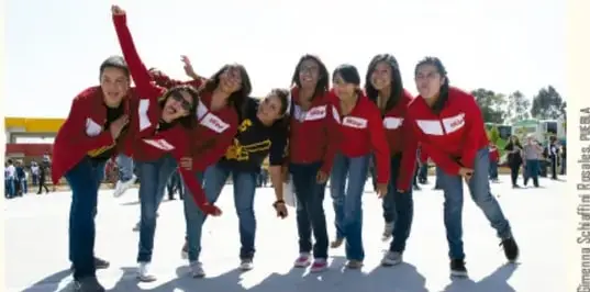 Fotografía de un grupo de jóvenes con playeras rojas que se agachan y sonríen posando juntos al aire libre; crédito de la fotógrafa en el lateral.