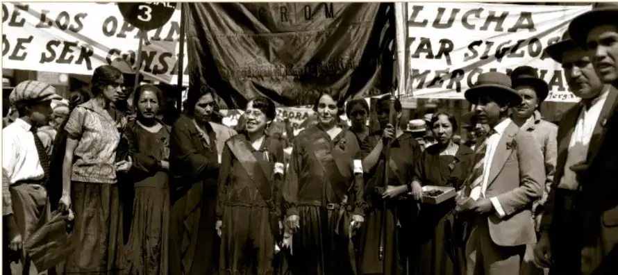 Fotografía histórica en sepia: mujeres y hombres de la Casa del Obrero Mundial se manifiestan con banderas y mantas con leyendas como 'LUCHA' y 'DE LOS OBREROS', exigiendo la igualdad civil y política. Crédito: Secretaría de Cultura, INAH, SINAFO, FN.MX. © (5884), 1924.