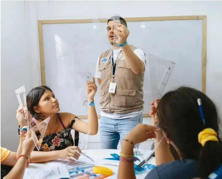 Docente y estudiantes de telesecundaria manipulando escuadras transparentes en el aula frente a un pizarrón.