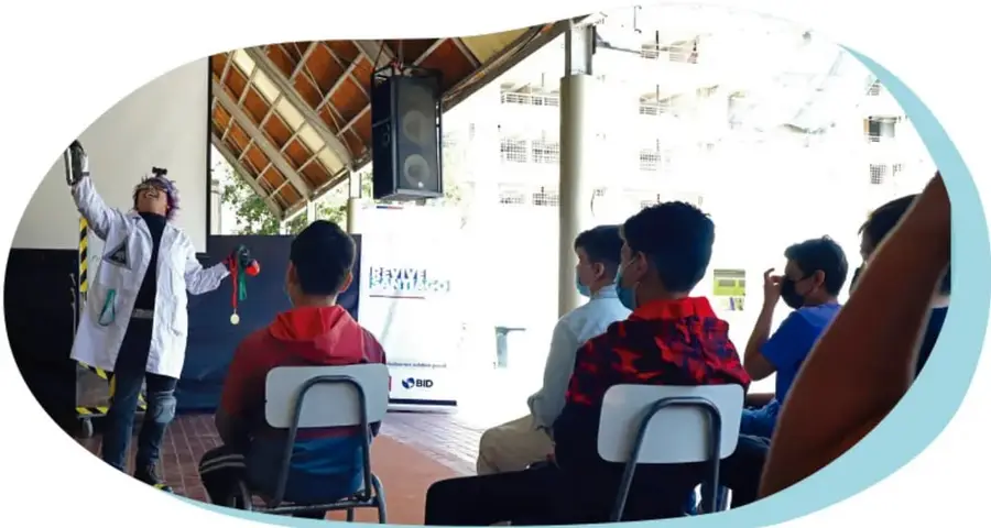 Fotografía en forma ovalada de una función teatral o presentación comunitaria: una persona disfrazada con bata blanca y gorro dirige la actividad frente a un público sentado en sillas, en un espacio techado al aire libre.