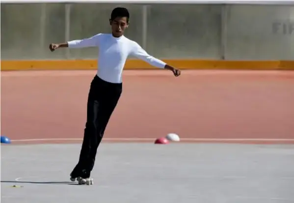 Fotografía de un patinador artístico adolescente con camisa blanca y pantalón negro, en pleno movimiento sobre una pista al aire libre.