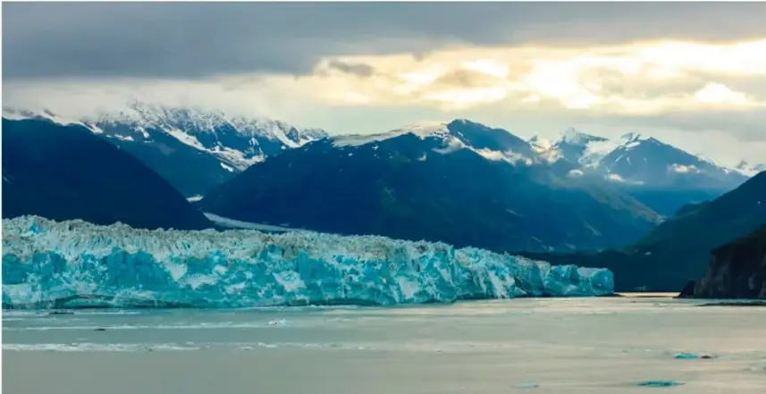 Fotografía de un glaciar azul en primer plano con montañas nevadas al fondo y un cielo con nubes iluminadas por el sol; ilustra la erosión glaciar y el modelado del relieve.