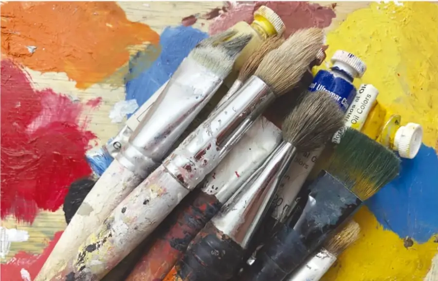 Fotografía de varios pinceles usados y tubos de pintura al óleo sobre una superficie con manchas de colores rojo, azul, amarillo y naranja.