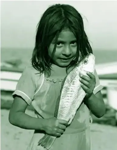 Fotografía en blanco y negro de una niña de cabello suelto, mirando al frente y sonriendo, que sostiene con ambas manos un pescado grande en posición vertical, cabeza hacia arriba; al fondo un paisaje costero desdibujado.