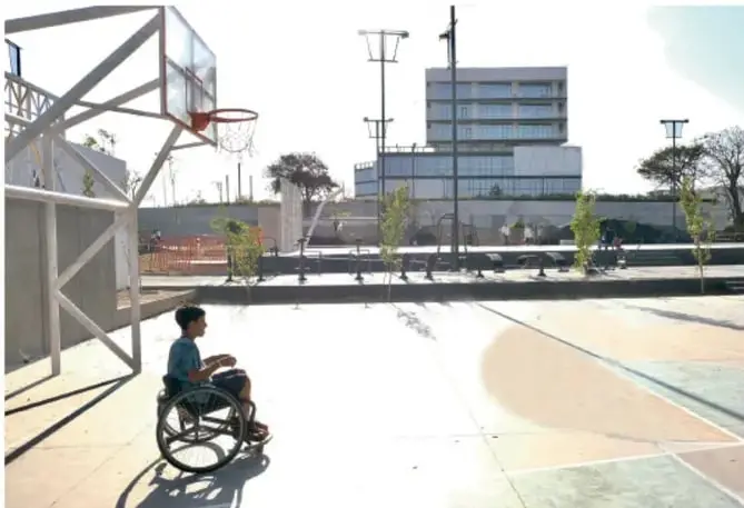 Persona en silla de ruedas en una cancha de básquetbol al aire libre, con estructuras metálicas y edificios al fondo.