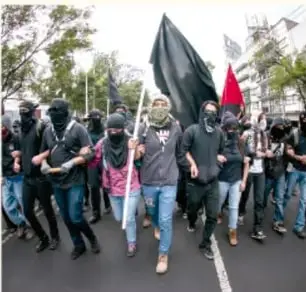Fotografía de una marcha con personas encapuchadas portando una bandera negra y una bandera roja.