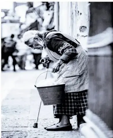 Fotografía en blanco y negro de una persona mayor inclinada lavando ropa en un barreño en la calle.
