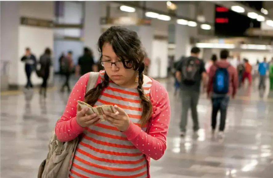 Fotografía de una estudiante leyendo un libro en un espacio público concurrido, posiblemente una estación o pasillo comercial.