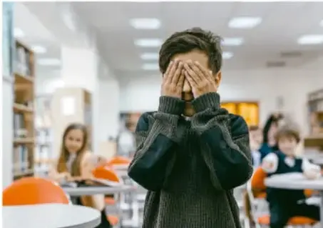 Fotografía de un niño con las manos en el rostro, en actitud de angustia, dentro de una biblioteca o aula con otros estudiantes al fondo.