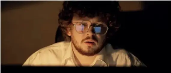 Fotografía de una persona con lentes retro iluminada por la pantalla de un equipo en un cuarto oscuro, representando el uso intensivo de la tecnología.