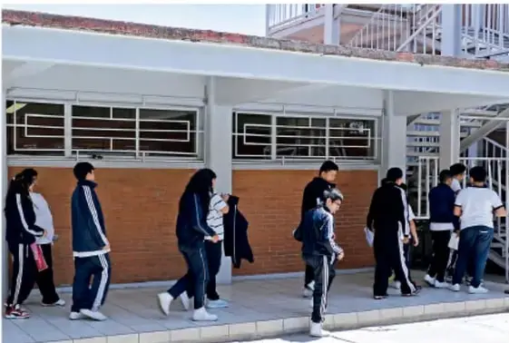 Fotografía de estudiantes de secundaria caminando en el patio o corredor de una escuela, algunos con cubrebocas, junto a un edificio escolar de ladrillo.