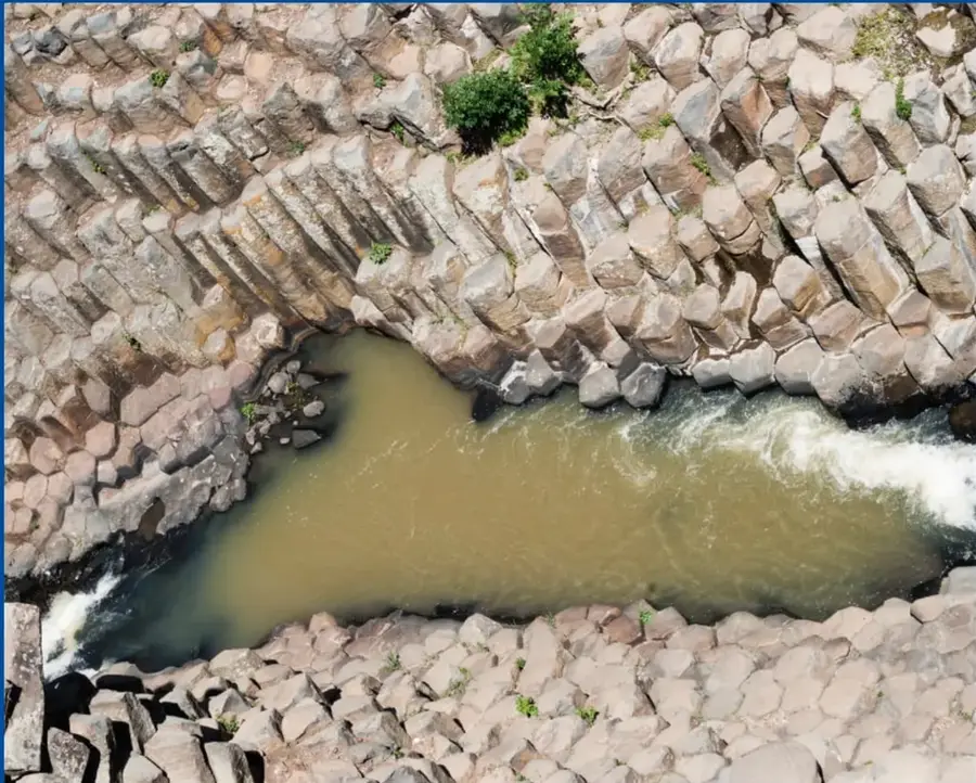 Fotografía aérea de un río de agua turbia y verdosa que fluye entre formaciones rocosas volcánicas de aspecto prismático (disyunción columnar), con pequeñas plantas creciendo en las grietas.
