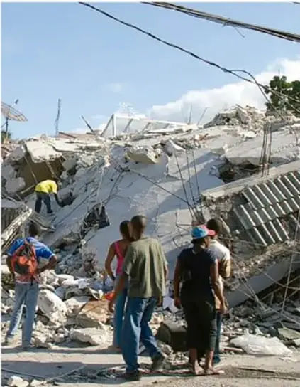 Fotografía de un grupo de personas caminando entre escombros de edificios derrumbados, con cielo despejado. Al fondo se observan estructuras colapsadas y cables eléctricos caídos.