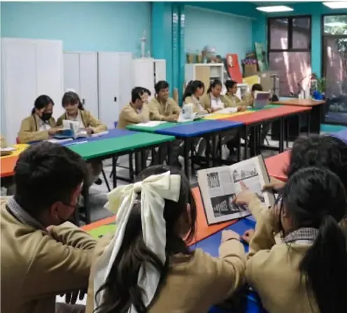 Grupo de estudiantes de telesecundaria trabajando en comunidad alrededor de mesas, revisando materiales y tablets, ilustrando el trabajo colaborativo de las etapas del proyecto.