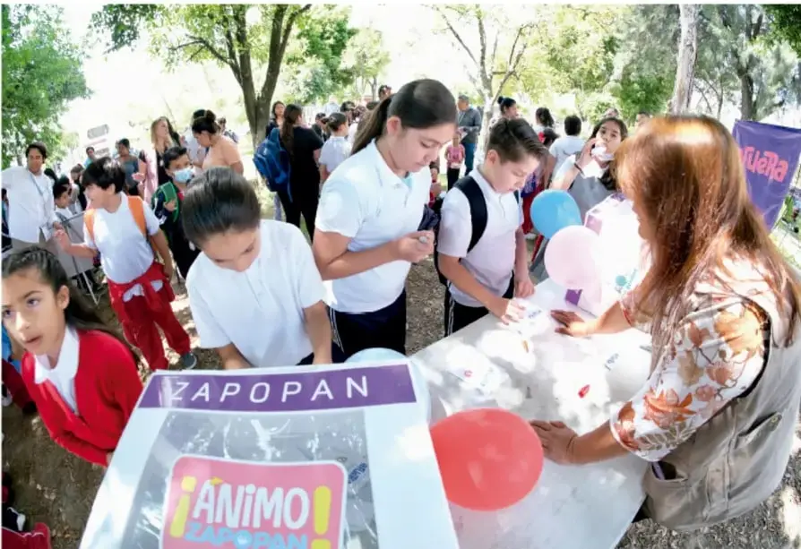 Estudiantes de primaria con uniforme participan en una exposición al aire libre; una persona adulta atiende un puesto con un cartel que dice ZAPOPAN y ÁNIMO ZAPOPAN, con globos y materiales sobre una mesa.