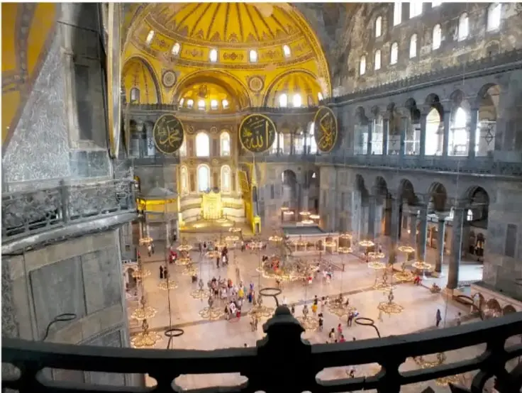 Fotografía interior de Santa Sofía en Estambul, antigua basílica bizantina convertida en mezquita, con gran cúpula dorada, mosaicos, medallones con caligrafía islámica y visitantes en la nave central.
