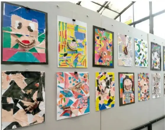 Muro escolar con varias obras plásticas colgadas: collages y pinturas coloridas con figuras y rostros.
