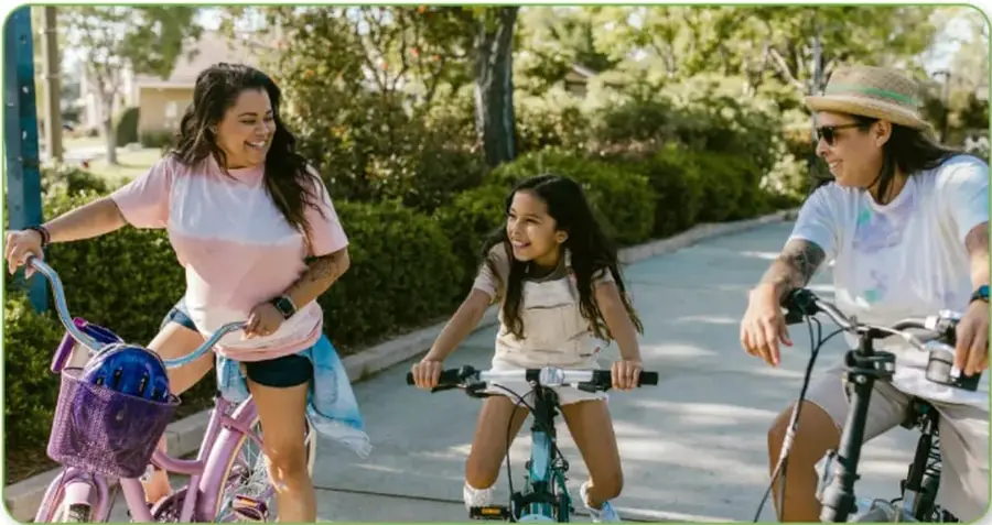 Fotografía de una familia en bicicleta en un parque: una mamá con camiseta rosa sobre una bici con canasta morada, una niña sonriente en su bici al centro y un papá con sombrero de palita y lentes a la derecha.