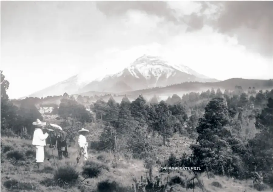Fotografía antigua en blanco y negro del Popocatépetl nevado; en primer plano, dos personas con sombrero junto a un burro entre arbustos y árboles.
